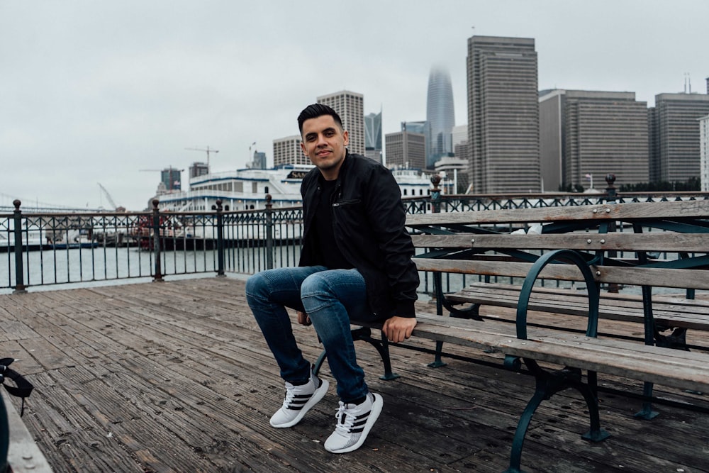 man in black jacket and blue denim jeans sitting on brown wooden bench