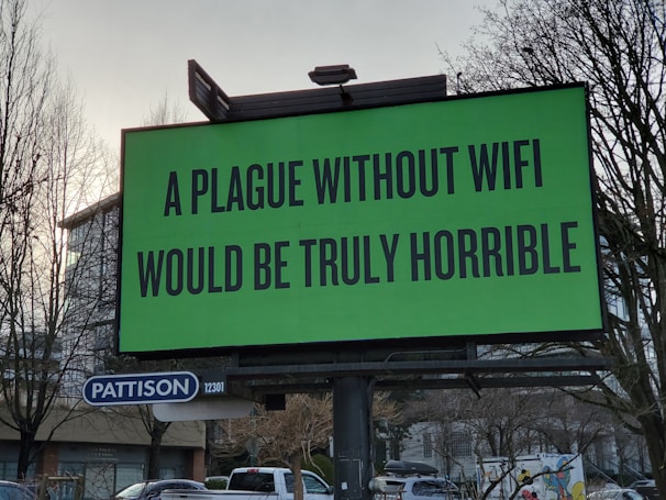 A large green billboard with the text 'A plague without WiFi would be truly horrible' in bold black font. Below the billboard is a smaller sign that says 'Pattison' with the number '12301'. Bare trees and a partially visible building are in the background.