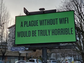 A large green billboard with the text 'A plague without WiFi would be truly horrible' in bold black font. Below the billboard is a smaller sign that says 'Pattison' with the number '12301'. Bare trees and a partially visible building are in the background.