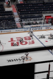 Ice hockey players are practicing on an indoor rink with empty spectator stands. The rink is marked with various logos, and the players are in full gear. The view is partially obstructed by a railing. Some players are standing, while others are kneeling or skating.