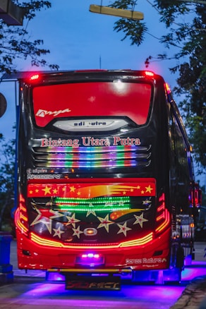 A brightly decorated bus with colorful LED lights and star patterns on the rear. The sky is a dusky blue, and the bus is parked or moving on a street lined with trees. The bus has a bold red and black color scheme with several brand names visible.