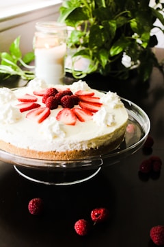 A cheesecake garnished with sliced strawberries and whole raspberries is placed on a glass cake stand. Whipped cream dollops are also arranged around the top of the cheesecake. In the background, there is a lit candle in a glass jar and a green leafy plant. Several raspberries are scattered on the dark surface around the cake stand.