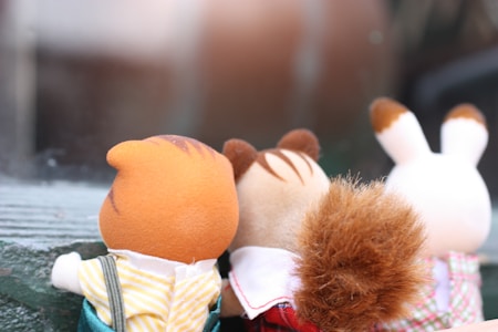 Three plush toy animals are positioned side by side with their backs facing the viewer. The toys include a brown and orange cat, a squirrel with a fluffy tail, and a white rabbit with brown ears. They appear to be looking out towards a window or a reflective surface.