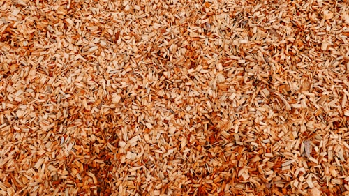 A close-up view of a large pile of wood chips, highlighting the texture and variety in size and shape of the fragments. The chips are mostly light brown with hints of darker tones.