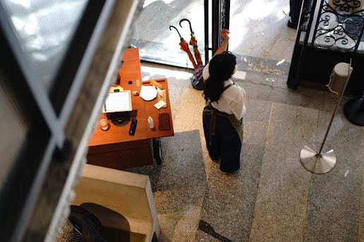A person wearing an apron and face mask stands in a room with a polished stone floor, near a small wooden desk. The desk holds various items including a tablet, a glass, and a bottle. Two umbrellas are propped near the entrance door that shows a decorative ironwork detail.