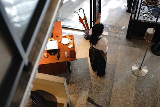 A person wearing an apron and face mask stands in a room with a polished stone floor, near a small wooden desk. The desk holds various items including a tablet, a glass, and a bottle. Two umbrellas are propped near the entrance door that shows a decorative ironwork detail.