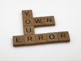 Wooden Scrabble tiles spell out the phrase 'OWN YOUR ERROR' arranged in a cross formation with letters in a light brown color against a white background.