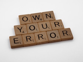 Wooden Scrabble tiles are arranged to spell out the phrase 'OWN YOUR ERROR' on a light surface.