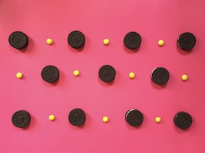 A series of black cookies and small yellow candies are arranged in a grid pattern on a vibrant pink background. The cookies are evenly spaced in straight lines, each interspersed with a yellow candy.