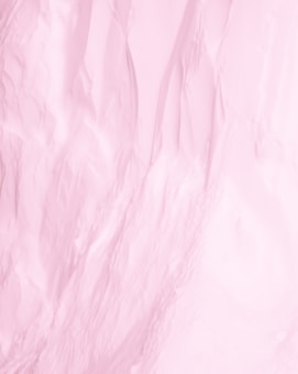 A close-up of a textured surface with soft folds, featuring a predominantly pale pink color. The surface appears to have a smooth, silky appearance reminiscent of fabric or a soft material.
