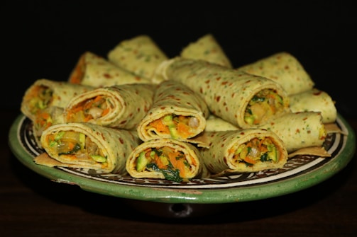 A beautifully arranged platter of rolled wraps filled with a colorful mix of vegetables such as carrots, zucchini, and spinach. The wraps are neatly placed on a decorative ceramic plate, creating an inviting and appetizing look.