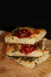Three slices of focaccia bread stacked on a wooden surface. The bread is garnished with herbs and roasted red peppers, creating a rustic and appetizing appearance.