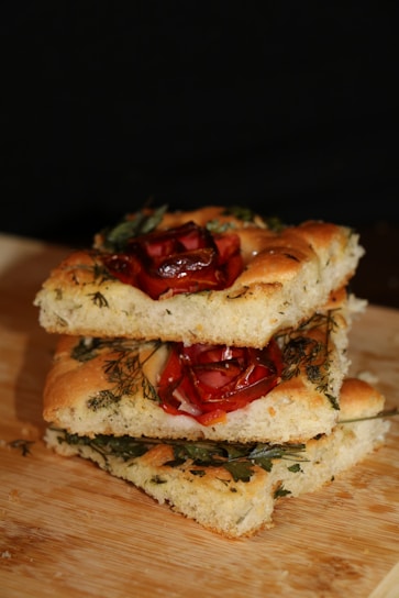 Three slices of focaccia bread stacked on a wooden surface. The bread is garnished with herbs and roasted red peppers, creating a rustic and appetizing appearance.