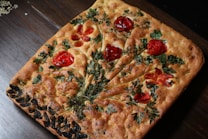 A beautifully baked focaccia bread topped with artistic placements of herbs and sliced tomatoes. The bread features a decorative pattern that resembles a bouquet of flowers, created with vibrant red tomatoes and green leaves. The crust is golden brown with a slightly rustic appearance.