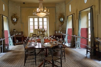 A vintage dining room with an elongated wooden table set with plates and cups, surrounded by antique chairs. The room features a large chandelier hanging from the ceiling, ornate mirrors on the walls, and draped windows allowing natural light to enter. Decor items such as vases and candelabras add a touch of elegance.