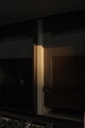 Dark and moody interior window with closed blinds casting shadows. A beam of warm light softly illuminates one section, adding a subtle contrast to the surrounding darkness.