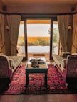 An elegant room with two vintage-style couches facing a glass door, leading to an outdoor terrace. A richly patterned red carpet covers the floor, and a wooden table is set with decorative items. The view outside shows a serene landscape with trees and a pool.