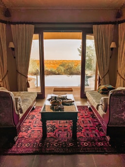 An elegant room with two vintage-style couches facing a glass door, leading to an outdoor terrace. A richly patterned red carpet covers the floor, and a wooden table is set with decorative items. The view outside shows a serene landscape with trees and a pool.