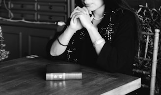 A monochrome image featuring a woman seated at a wooden table, her hands clasped near her chin. She wears a dark outfit and a pearl necklace, with a book placed on the table in front of her. The background includes a set of drawers and some foliage.