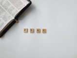 An open book with Chinese text lies beside four beige Scrabble tiles spelling the word 'PRAY' on a white surface.
