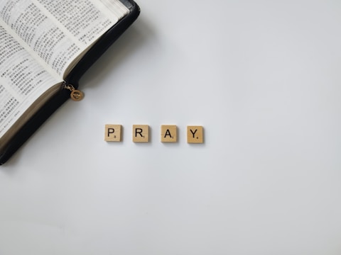 An open book with Chinese text lies beside four beige Scrabble tiles spelling the word 'PRAY' on a white surface.