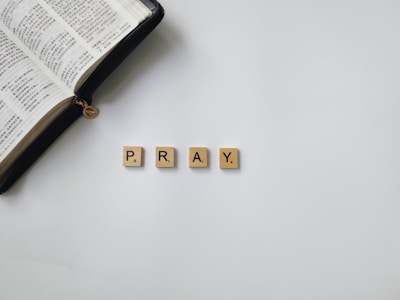An open book with Chinese text lies beside four beige Scrabble tiles spelling the word 'PRAY' on a white surface.