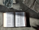 An open book with text in a language that appears to be Chinese is placed on a textured surface. There is a glass of water nearby, showing light reflections and refractions. Part of a newspaper with visible text is placed adjacent to the book, adding a sense of casual reading or study.