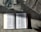 An open book with text in a language that appears to be Chinese is placed on a textured surface. There is a glass of water nearby, showing light reflections and refractions. Part of a newspaper with visible text is placed adjacent to the book, adding a sense of casual reading or study.