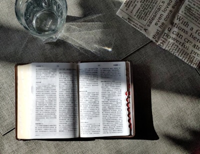 An open book with text in a language that appears to be Chinese is placed on a textured surface. There is a glass of water nearby, showing light reflections and refractions. Part of a newspaper with visible text is placed adjacent to the book, adding a sense of casual reading or study.