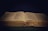 An open book rests against a dark background, the pages filled with small, printed text. The book's cover is textured with a golden hue, and a thin ribbon bookmark lies in the center.