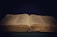 An open book rests against a dark background, the pages filled with small, printed text. The book's cover is textured with a golden hue, and a thin ribbon bookmark lies in the center.