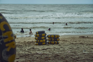A group of inner tubes with yellow lettering and designs are stacked on a sandy beach. In the background, several people are swimming in the ocean, with waves visible in the water.