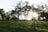 A serene park setting with groups of people sitting on the grass, enjoying the late afternoon sun. The sunlight filters through the trees, creating long shadows on the ground. In the background, a building partially obscured by the foliage can be seen.