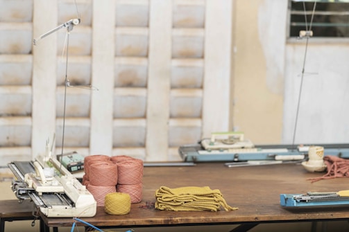 A wooden table holds several spools of yarn in pink and yellow colors, along with pieces of knitted fabric. Knitting machines are positioned on the table, and the background features partially blurred walls with a textured pattern.