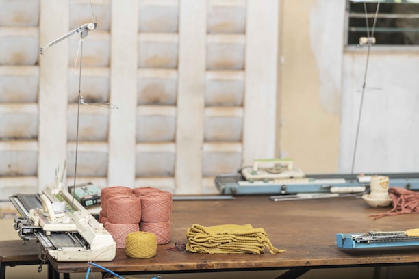 A wooden table holds several spools of yarn in pink and yellow colors, along with pieces of knitted fabric. Knitting machines are positioned on the table, and the background features partially blurred walls with a textured pattern.