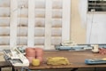 A wooden table holds several spools of yarn in pink and yellow colors, along with pieces of knitted fabric. Knitting machines are positioned on the table, and the background features partially blurred walls with a textured pattern.