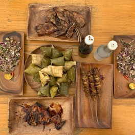 A wooden table is set with several wooden plates containing different dishes. At the center, a circular plate holds woven rice packages known as 'puso'. Surrounding it are plates with grilled meats, chopped dishes with visible onions and green herbs, and two bottles of dark and clear liquid. The arrangement suggests a traditional or cultural meal setting.