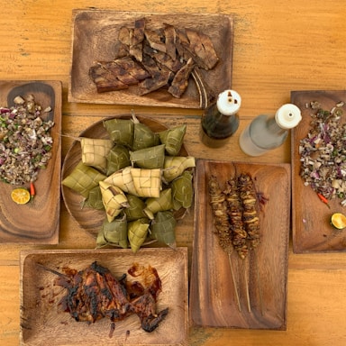 A wooden table is set with several wooden plates containing different dishes. At the center, a circular plate holds woven rice packages known as 'puso'. Surrounding it are plates with grilled meats, chopped dishes with visible onions and green herbs, and two bottles of dark and clear liquid. The arrangement suggests a traditional or cultural meal setting.