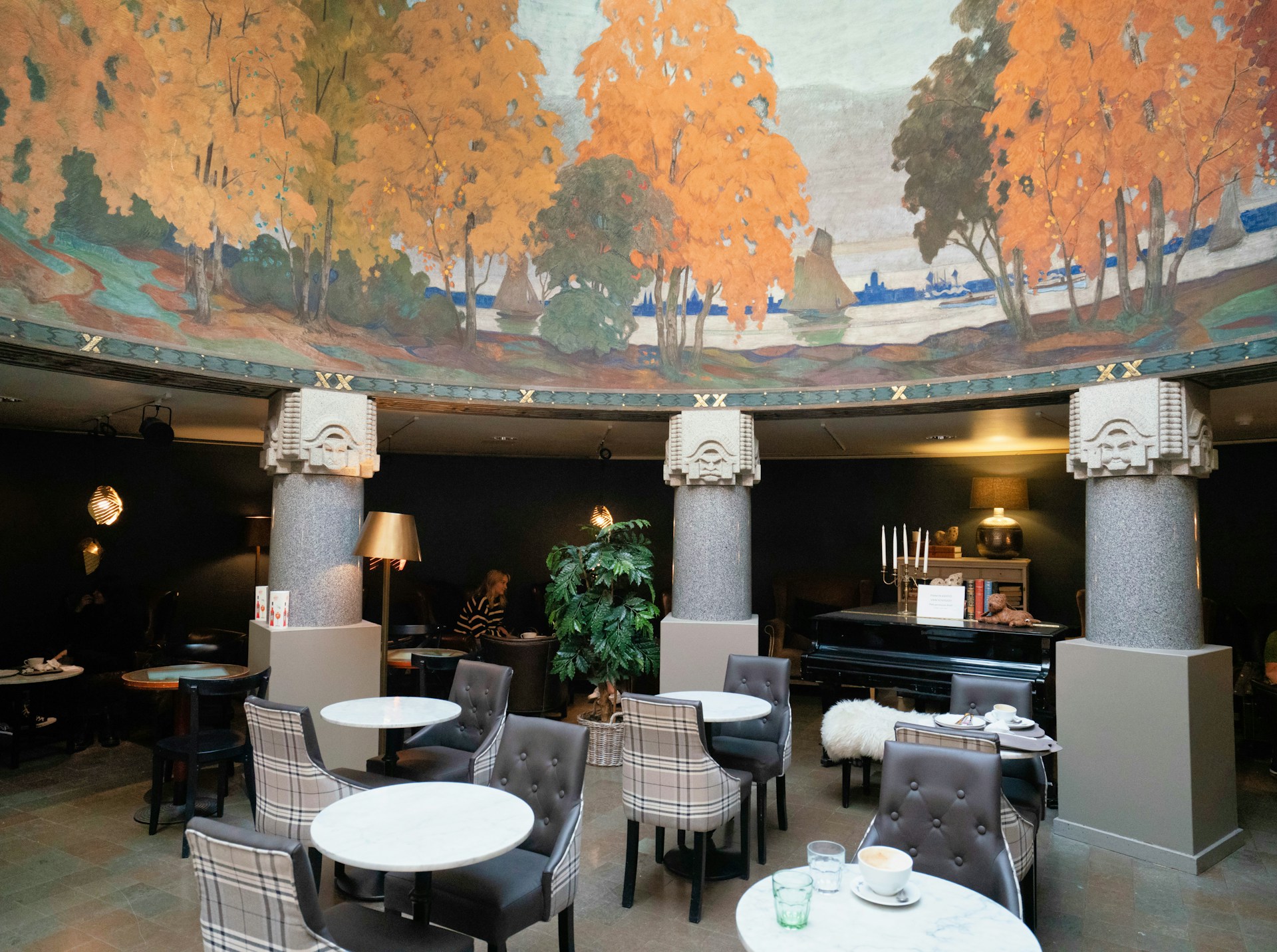 A cozy café interior featuring elegant columns and round tables with upholstered chairs. The ceiling displays a large mural depicting a colorful autumn landscape with vibrant orange and green trees. Soft lighting and a grand piano enhance the sophisticated atmosphere.