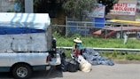 A small truck with a makeshift covering is parked on the side of the street near a collection of trash bags. A person wearing a straw hat and colorful clothing is standing next to the bags. Behind them is a wire fence and a commercial area with trailers and advertising signs in Spanish.