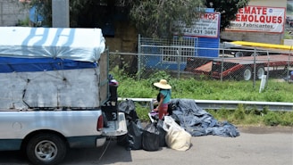 A small truck with a makeshift covering is parked on the side of the street near a collection of trash bags. A person wearing a straw hat and colorful clothing is standing next to the bags. Behind them is a wire fence and a commercial area with trailers and advertising signs in Spanish.
