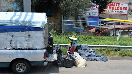 A small truck with a makeshift covering is parked on the side of the street near a collection of trash bags. A person wearing a straw hat and colorful clothing is standing next to the bags. Behind them is a wire fence and a commercial area with trailers and advertising signs in Spanish.