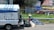 A small truck with a makeshift covering is parked on the side of the street near a collection of trash bags. A person wearing a straw hat and colorful clothing is standing next to the bags. Behind them is a wire fence and a commercial area with trailers and advertising signs in Spanish.
