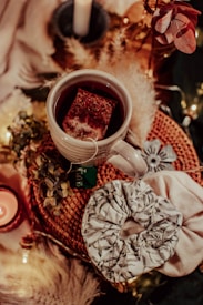 A cozy arrangement features a white mug of tea with a teabag inside, sitting on a woven mat. Nearby, there are decorative elements such as dried flowers, a lit candle, and a scrunchie with a leafy pattern. Soft lighting and warm colors create an inviting atmosphere.
