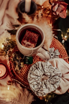 A cozy arrangement features a white mug of tea with a teabag inside, sitting on a woven mat. Nearby, there are decorative elements such as dried flowers, a lit candle, and a scrunchie with a leafy pattern. Soft lighting and warm colors create an inviting atmosphere.