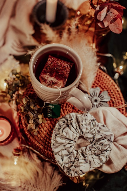 A cozy arrangement features a white mug of tea with a teabag inside, sitting on a woven mat. Nearby, there are decorative elements such as dried flowers, a lit candle, and a scrunchie with a leafy pattern. Soft lighting and warm colors create an inviting atmosphere.