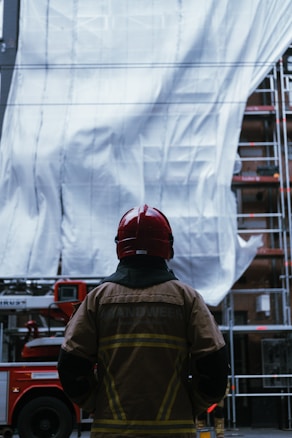 A person wearing a red helmet and a fire jacket stands in front of a large backdrop, possibly a building or construction site. A red fire truck is partially visible behind the person, along with metal scaffolding.