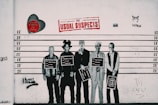 A lineup of five well-known figures depicted as if in a police lineup, each holding a sign with their name and artistic movement. The background includes various graffiti elements, a red heart with text, and a large 'The Usual Suspects' sign in red lettering. Two more pieces of graffiti include a stylized cat and the words 'Almost Criminal' sprayed in black.