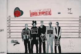 A lineup of five well-known figures depicted as if in a police lineup, each holding a sign with their name and artistic movement. The background includes various graffiti elements, a red heart with text, and a large 'The Usual Suspects' sign in red lettering. Two more pieces of graffiti include a stylized cat and the words 'Almost Criminal' sprayed in black.