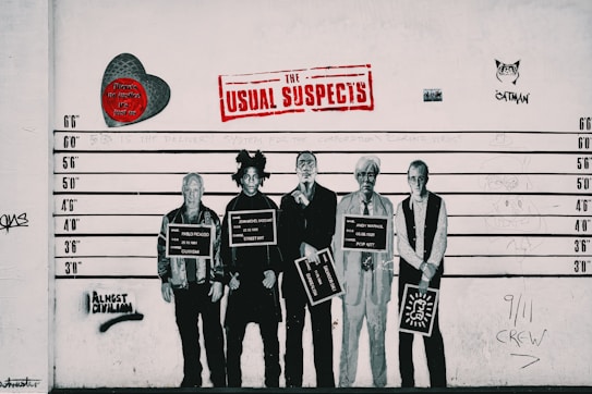 A lineup of five well-known figures depicted as if in a police lineup, each holding a sign with their name and artistic movement. The background includes various graffiti elements, a red heart with text, and a large 'The Usual Suspects' sign in red lettering. Two more pieces of graffiti include a stylized cat and the words 'Almost Criminal' sprayed in black.